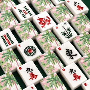 May include: A collection of white Mahjong tiles with red, green, and black symbols and numbers. Some tiles feature a pink and green palm tree design. The tiles are arranged on a dark green surface.