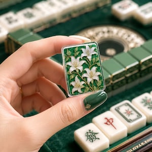 Pu&ograve; includere: Una tessera rettangolare da mahjong con uno sfondo verde vivace decorato da un'illustrazione dettagliata di tre fiori di giglio bianchi con steli e foglie verdi, racchiusa in un bordo protettivo trasparente per una finitura liscia.