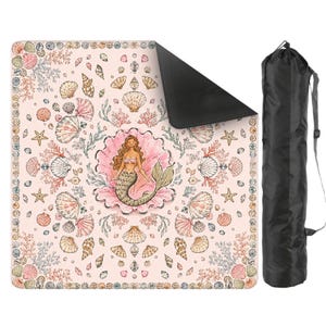 May include: A square picnic blanket with a light pink background featuring a central illustration of a mermaid inside a large seashell, surrounded by various seashells, starfish, and coral patterns, accompanied by a black cylindrical carrying bag.