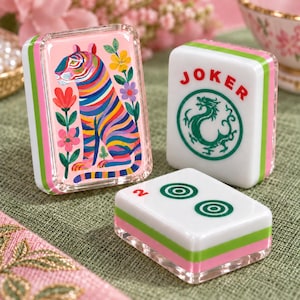 May include: Three rectangular Mahjong tiles with layered pink, green, and white edges. One tile features a colorful tiger and floral illustration. Another tile displays the red text JOKER above a green dragon icon. The third tile shows a red number 2.