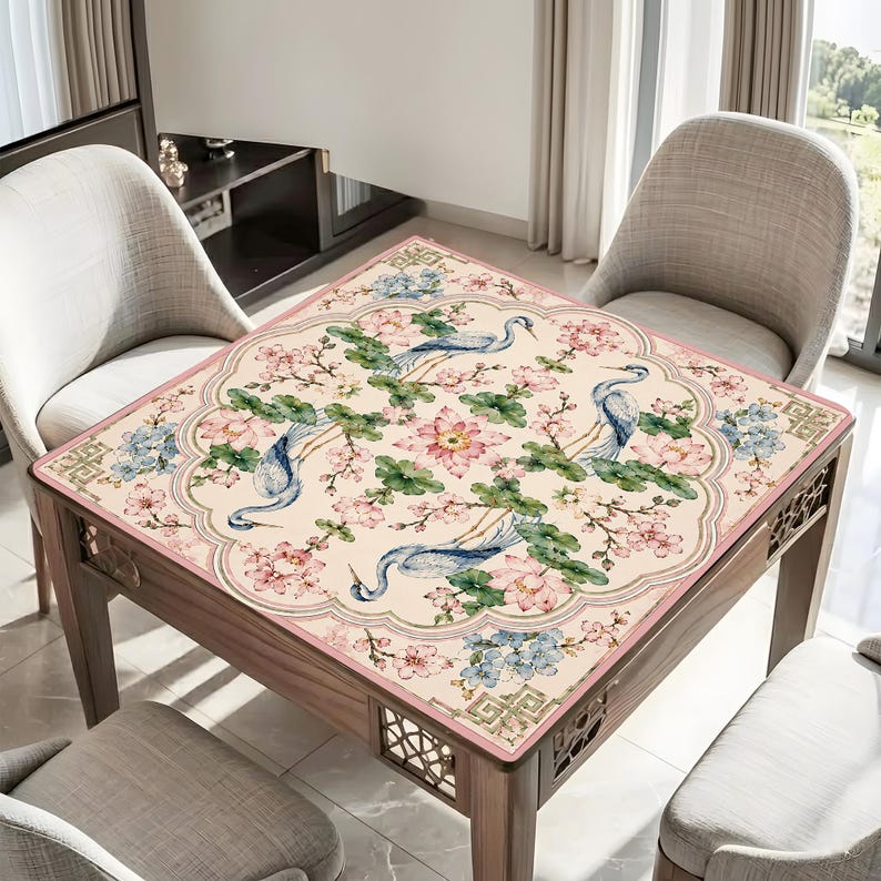 May include: A square wooden table with a floral patterned top featuring blue herons, pink flowers, and green leaves. The table has a light pink border and is surrounded by chairs.