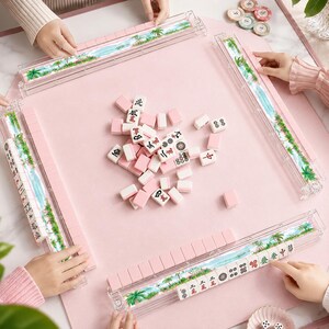 May include: A pink Mahjong set with white and pink tiles, and clear plastic tile holders with a tropical landscape design. The set is arranged on a pink surface, with hands visible around the table.