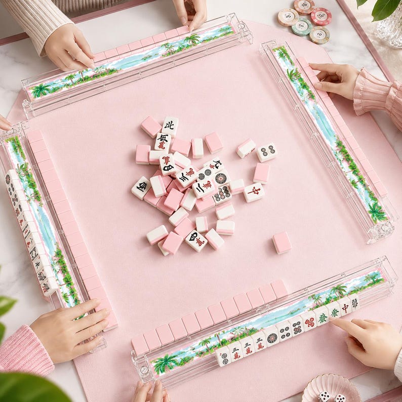 May include: A pink Mahjong set with white and pink tiles, and clear plastic tile holders with a tropical landscape design. The set is arranged on a pink surface, with hands visible around the table.