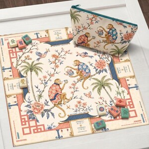 Op de afbeelding: Een vierkante Mahjong-mat en een bijpassend tasje met rits op een crèmekleurige achtergrond, voorzien van illustraties van apen in patronen, palmbomen, bloemen en vogels. Diverse kleurrijke Mahjong-stenen liggen verspreid op de mat.
