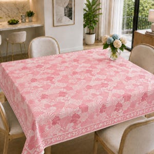 May include: A square tablecloth featuring a repeating pattern of white seashells on a soft pink background. The fabric has a distinct border design with a continuous seashell motif along the edges, covering a dining table with four chairs.