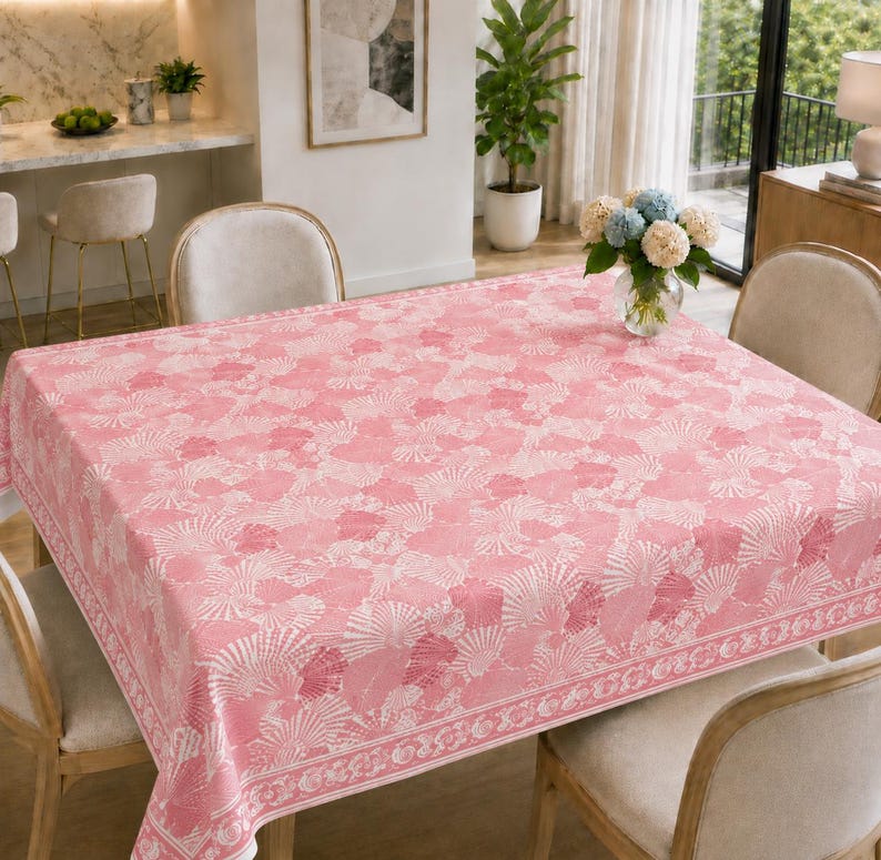 May include: A square tablecloth featuring a repeating pattern of white seashells on a soft pink background. The fabric has a distinct border design with a continuous seashell motif along the edges, covering a dining table with four chairs.