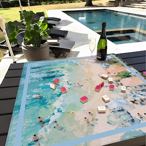 May include: A Mahjong set with a beach scene on the board. The board shows an ocean with swimmers. Pink and white tiles are scattered on the board. A bottle of champagne and a glass are also on the table.
