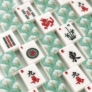 May include: A collection of white Mahjong tiles with red, green, and black symbols and characters. The tiles are arranged on a patterned background with a floral design in shades of teal and white. Some tiles feature a red dragon.