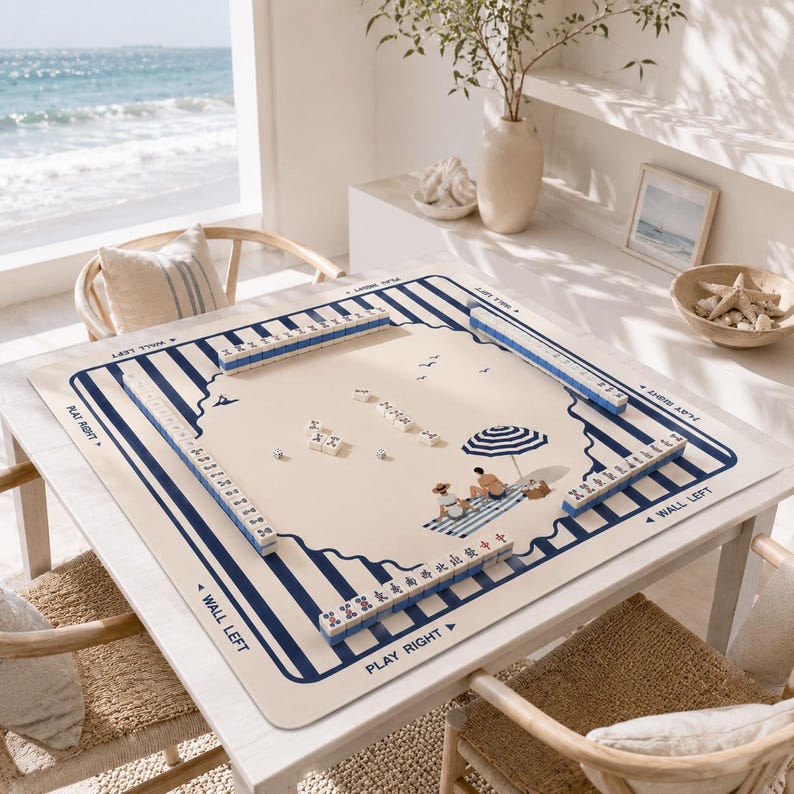 May include: A mahjong set with blue and white tiles and a beige playing surface. The surface features a beach scene with an umbrella and figures. The set is on a white table with a view of the ocean.