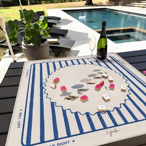 May include: A beige and blue striped Mahjong mat with a wavy border, featuring a scattered game setup with tiles and miniature beach scenes. A bottle of champagne and a glass are on the table. The scene is set outdoors near a pool.