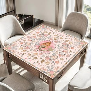 May include: A square table cover featuring a light pink background with a central illustration of a mermaid resting inside a large seashell. The surface is decorated with a repeating pattern of various seashells, starfish, and coral in soft tones.