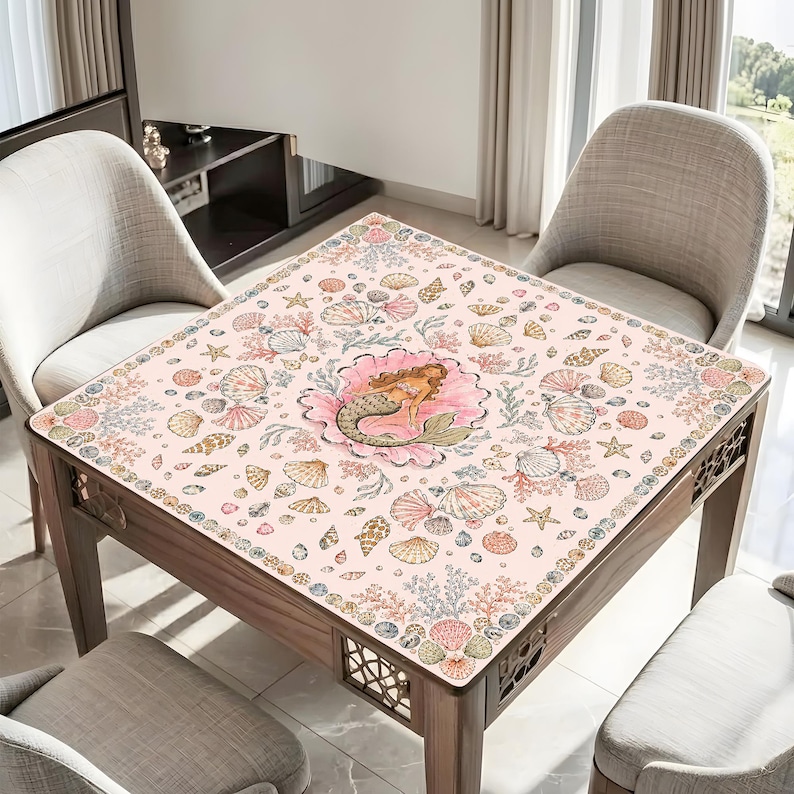 May include: A square table cover featuring a light pink background with a central illustration of a mermaid resting inside a large seashell. The surface is decorated with a repeating pattern of various seashells, starfish, and coral in soft tones.