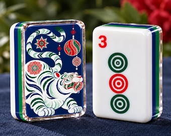 May include: Two rectangular mahjong tiles with rounded edges. One tile features a dark blue background with a white tiger illustration, red and green floral accents, and hanging ornaments. The other white tile displays a red number 3 and three circles.