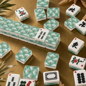 May include: A collection of Mahjong tiles with a floral design in shades of green and white. The tiles are arranged on a gold-colored surface. Some tiles have Chinese characters and numbers.