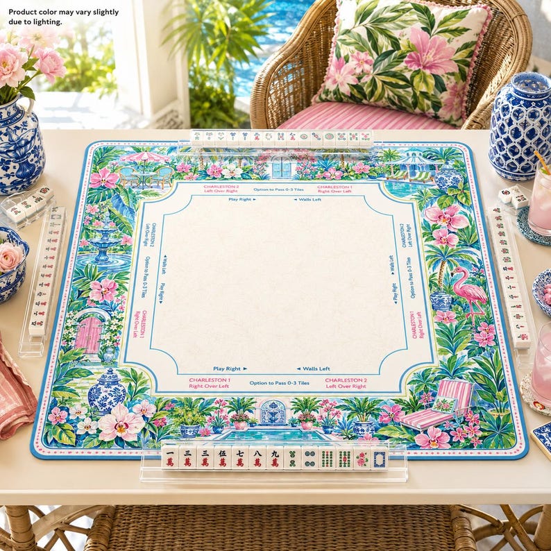 May include: A square mahjong mat featuring a vibrant tropical garden border with pink flowers, green palm leaves, a flamingo, and blue fountains. The center is cream-colored with printed game instructions and directional arrows in blue and pink text.