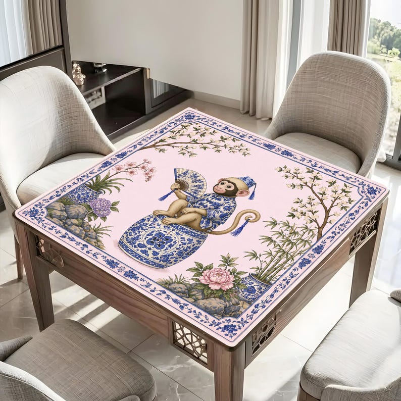 May include: A square table with a pink surface featuring a blue and white illustration of a monkey in a decorative vase, holding a fan. The table has a wooden frame and is surrounded by chairs.