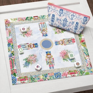 May include: A square game mat featuring a watercolor print of nutcrackers, pink poinsettias, and a Greek key border, accompanied by a matching zippered pouch with a blue and white chinoiserie pattern and a pink zipper, along with five game tiles.