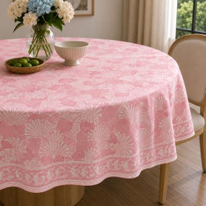May include: A round pink tablecloth featuring a white seashell and coral pattern with a decorative border. The fabric drapes over a circular table, providing a soft color palette for the dining area while maintaining a clean and organized appearance.