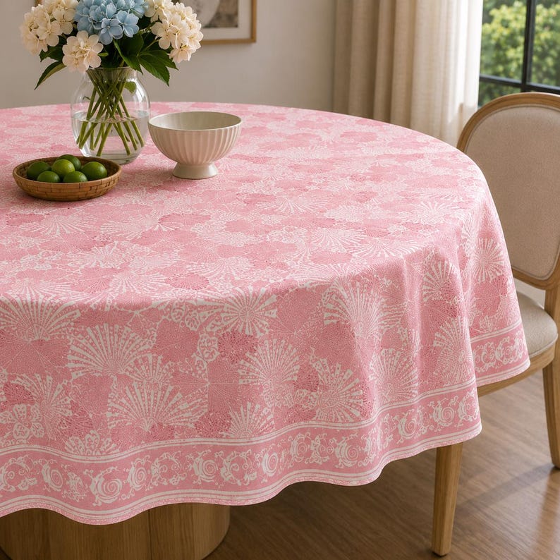May include: A round pink tablecloth featuring a white seashell and coral pattern with a decorative border. The fabric drapes over a circular table, providing a soft color palette for the dining area while maintaining a clean and organized appearance.