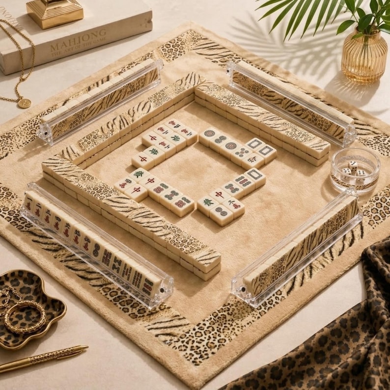 May include: A mahjong set featuring cream-colored tiles with black, red, and green symbols, arranged on a tan mat with a leopard and tiger print border. The set includes four clear acrylic tile racks and a small clear bowl containing dice.