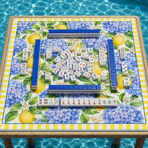 May include: A square Mahjong table with a floral and lemon print, set outdoors. The table features blue and white tiles, arranged for play. The table's edge has a yellow and white striped border.