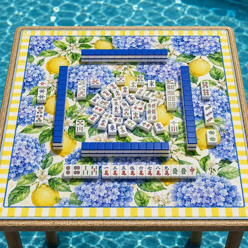May include: A square Mahjong table with a floral and lemon print, set outdoors. The table features blue and white tiles, arranged for play. The table's edge has a yellow and white striped border.