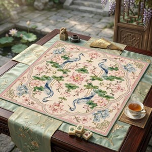 May include: A decorative square mat with a floral and bird design. The mat features blue herons, pink and blue flowers, and green leaves on a cream background, framed by a pink border. The mat is placed on a dark wooden table with green runners.