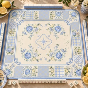 May include: A square mahjong game mat with a floral design in shades of blue and cream. The mat features a central floral motif and decorative borders with plaid and floral patterns. The set includes tiles and racks, creating a cohesive game setup.