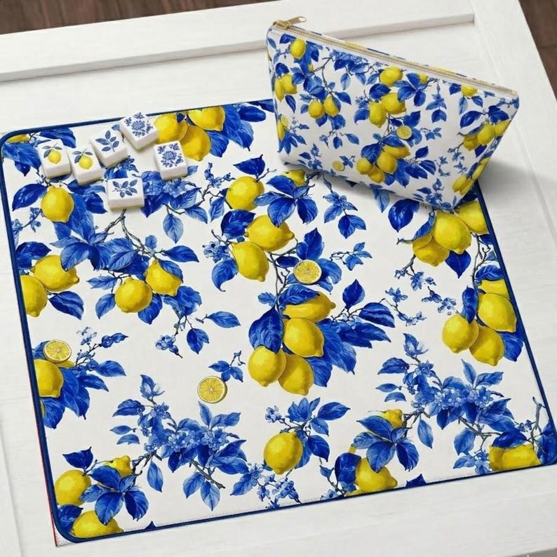 May include: A white mat and cosmetic bag with a lemon and blue floral design. The mat has a blue border and is covered in yellow lemons and blue leaves. The cosmetic bag has a gold zipper. Several decorative tiles are on the mat.