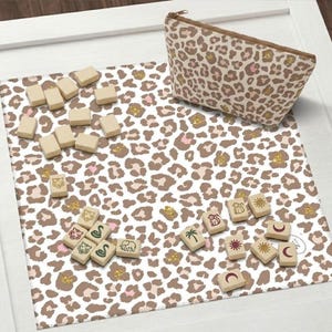 Könnte beinhalten: Eine Kosmetiktasche mit Leopardenmuster, eine passende Matte und Holzspielsteine. Die Matte und die Tasche weisen ein braunes, rosa und goldenes Leopardenmuster auf. Die Spielsteine haben verschiedene Tier- und Symbolillustrationen.