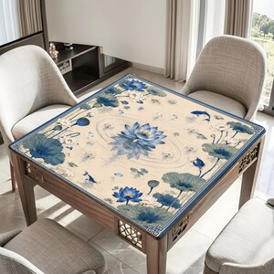 May include: A square table cover featuring a cream-colored background with a blue lotus flower and lily pad pattern. The design includes circular ripples, small fish, and a decorative blue geometric border around the edges of the fabric.