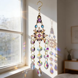 Puede incluir: Un atrapasoles decorativo con un marco de metal dorado y cristales colgantes. El atrapasoles presenta un diseño de estrella con cristales morados, azules y transparentes. Cuelga de un gancho dorado.