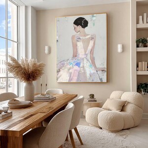 Puede incluir: Pintura abstracta de una mujer con un vestido blanco con la espalda baja, enmarcada en oro. La obra de arte cuelga sobre una mesa de comedor de madera con sillas beige. Un sill&oacute;n esponjoso de color crema y un jarr&oacute;n con hierba de la pampa seca completan la decoraci&oacute;n de la habitaci&oacute;n.