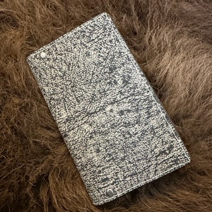 May include: A rectangular, leather-bound journal with a speckled black and cream pattern. The journal is closed and secured with two silver rivets. The background is a textured, brown surface.
