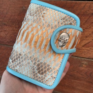 May include: A light blue leather wallet with a gold and white snakeskin pattern. The wallet has a skull-shaped button closure.