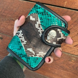 May include: A green and brown snakeskin wallet with a black leather trim and a silver skull accent.