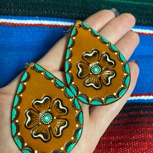 Op de afbeelding: Een paar druppelvormige oorbellen. De oorbellen zijn bruin met een turquoise rand en een bloemmotief in het midden. Het bloemmotief is bruin en wit met een turquoise centrum. De oorbellen worden in een hand gehouden.