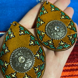 Op de afbeelding: Een paar druppelvormige oorbellen in mosterdgeel met een western thema. De oorbellen hebben een zilverkleurig bloemendesign in het midden en turquoise en gele accenten. De oorbellen worden in een hand gehouden.