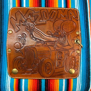 Op de afbeelding: Een rechthoekig bruin lederen paneel met een gedetailleerde reliëfafbeelding van een cartoonmuis die in een hot rod rijdt. De tekst MAZDOOMA! staat bovenaan in het leer gedrukt. Het leer is bevestigd met vier koperkleurige metalen klinknagels.