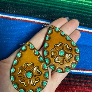 Op de afbeelding: Een paar druppelvormige oorbellen met een bruine leerlook. Elke oorbel heeft een centraal bloemmotief en een rand van turquoise en crèmekleurige accenten. De oorbellen worden in een hand gehouden.