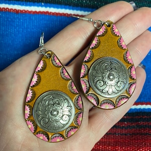 Op de afbeelding: Een paar druppelvormige oorbellen met een western thema. De oorbellen hebben een zilverkleurig medaillon met bloemendetails, geplaatst op een bruine leren basis met roze en gele stippen. De oorbellen hangen aan zilveren haken.