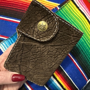 May include: A brown leather wallet with a skull and crossbones button. The wallet has a textured surface and is folded in half.