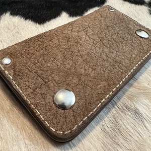 Op de afbeelding: Een bruine leren portemonnee met witte stiksels en zilveren klinknagels. De portemonnee is gesloten en ligt op een wit en zwart koeienhuid oppervlak.