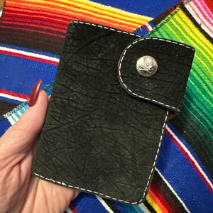 May include: A black leather wallet with a skull and crossbones button closure. The wallet has a textured surface and white stitching.