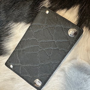 May include: A black leather wallet with a textured surface and silver accents. The wallet has a unique design and is made of high-quality materials.