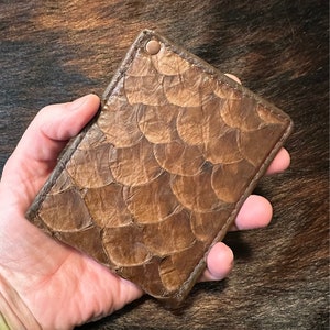 May include: A brown leather wallet with a unique, textured, fish-scale pattern. The wallet has a single, copper-colored rivet on the top right corner.