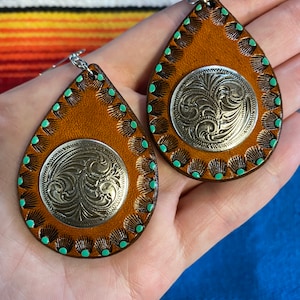 Op de afbeelding: Een paar druppelvormige oorbellen. De oorbellen zijn gemaakt van bruin leer met een zilverkleurige, cirkelvormige versiering in het midden. De randen zijn versierd met turquoise accenten. De oorbellen hebben zilverkleurige haken.