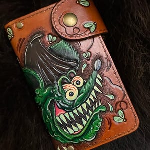 May include: A rectangular brown leather wallet featuring a hand-tooled and painted graphic of a green, sharp-toothed creature with bulging eyes and a dark wing. The wallet includes a snap closure, brass-toned hardware, and small green fly accents.
