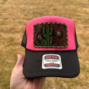 May include: A bright pink and black trucker hat featuring a rectangular brown leather patch with an embossed green cactus design and turquoise stitching. The black brim displays a white sticker with the text OTTO WORLD FAMOUS FOAM FRONT.