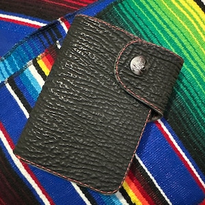 May include: A black leather wallet with a skull and crossbones button closure. The wallet is lying on a colorful striped blanket.